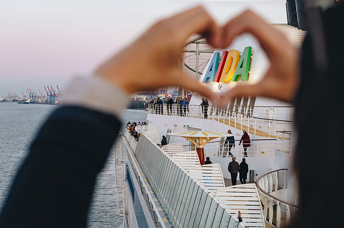 unscharfes mit den Haenden geformtes Herz; im Hintergrund scharfgestellt ein AIDA-Kreuzfahrtschiff; das AIDA-Logo des Schiffes ist genau in dem Herz